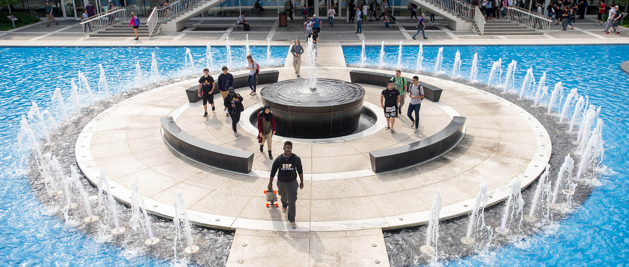 Students walk through UAlbany's academic podium, which includes staircases, overhangs, benches and a fountain.