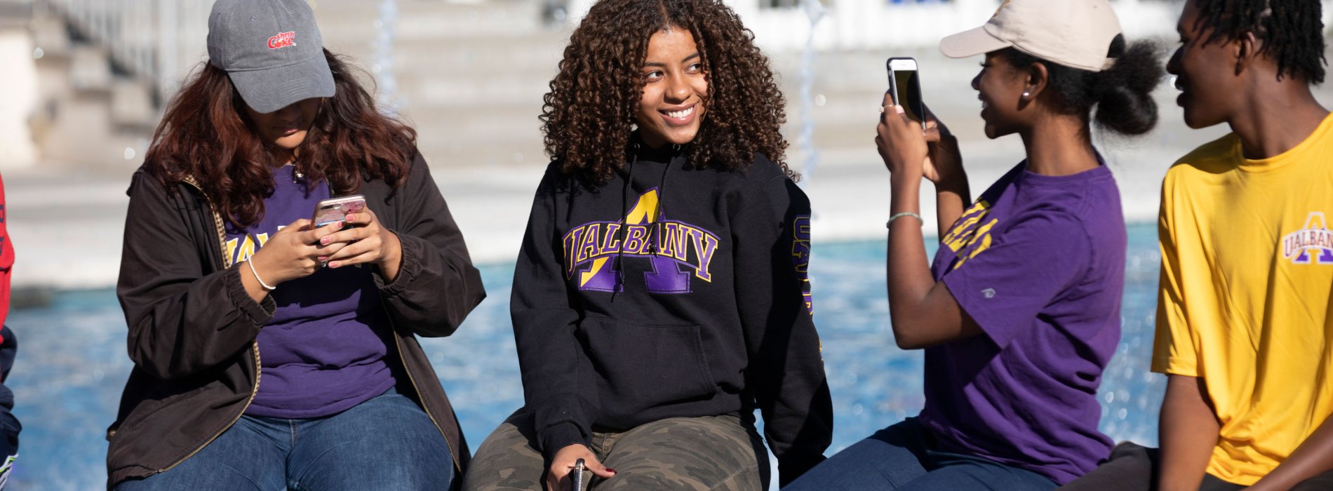 Four students sit on the edge of a fountain, one poses for a photo that the other is taking.